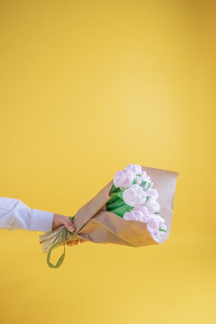 A person in a white shirt holds a bouquet of fresh, pastel pink tulips wrapped in light brown kraft paper with a green satin ribbon, presented against a vibrant yellow background. The tulips have smoo